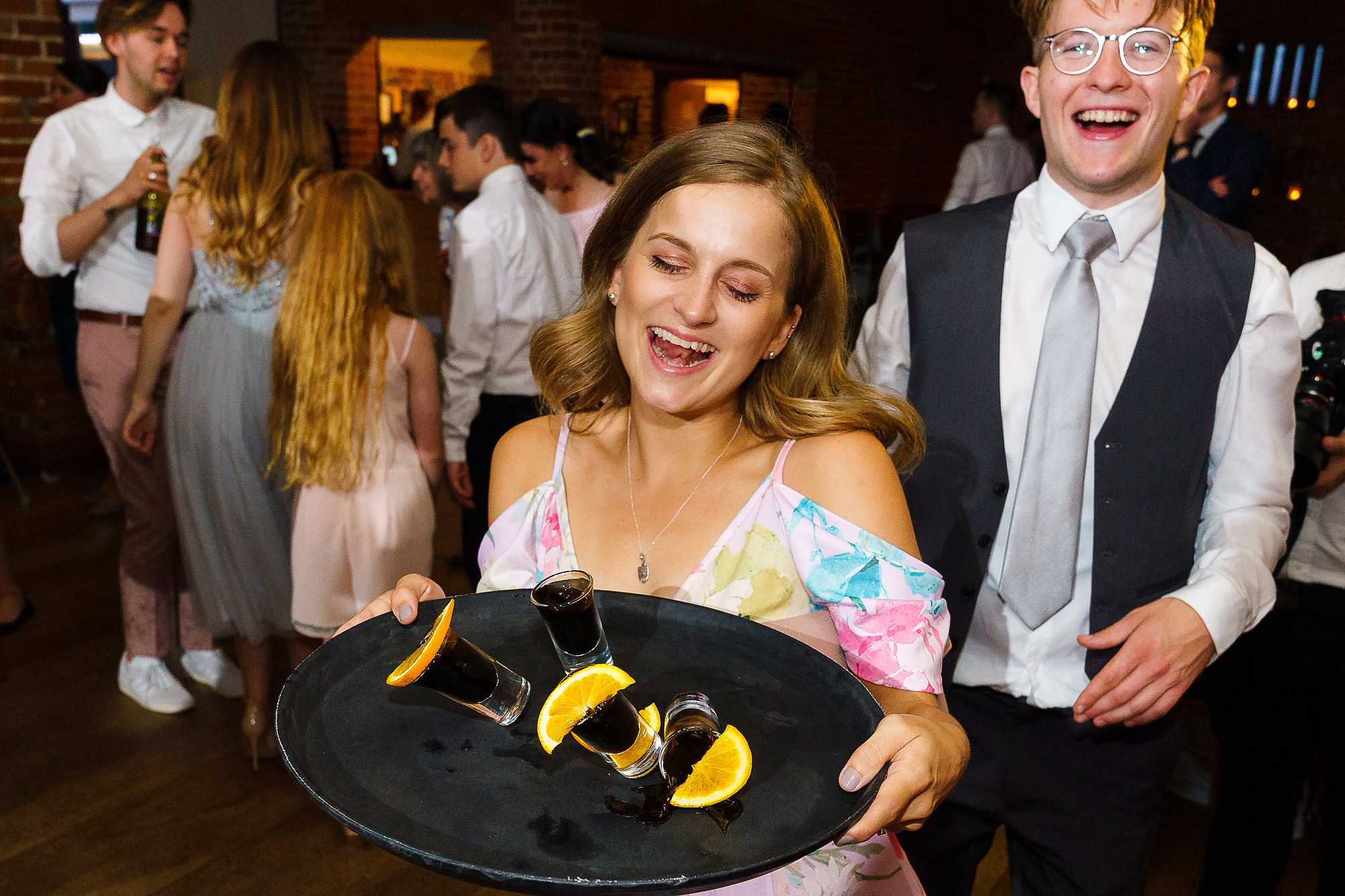 Woman with tray of shots and orange slices at a social gathering, lively party in background.
