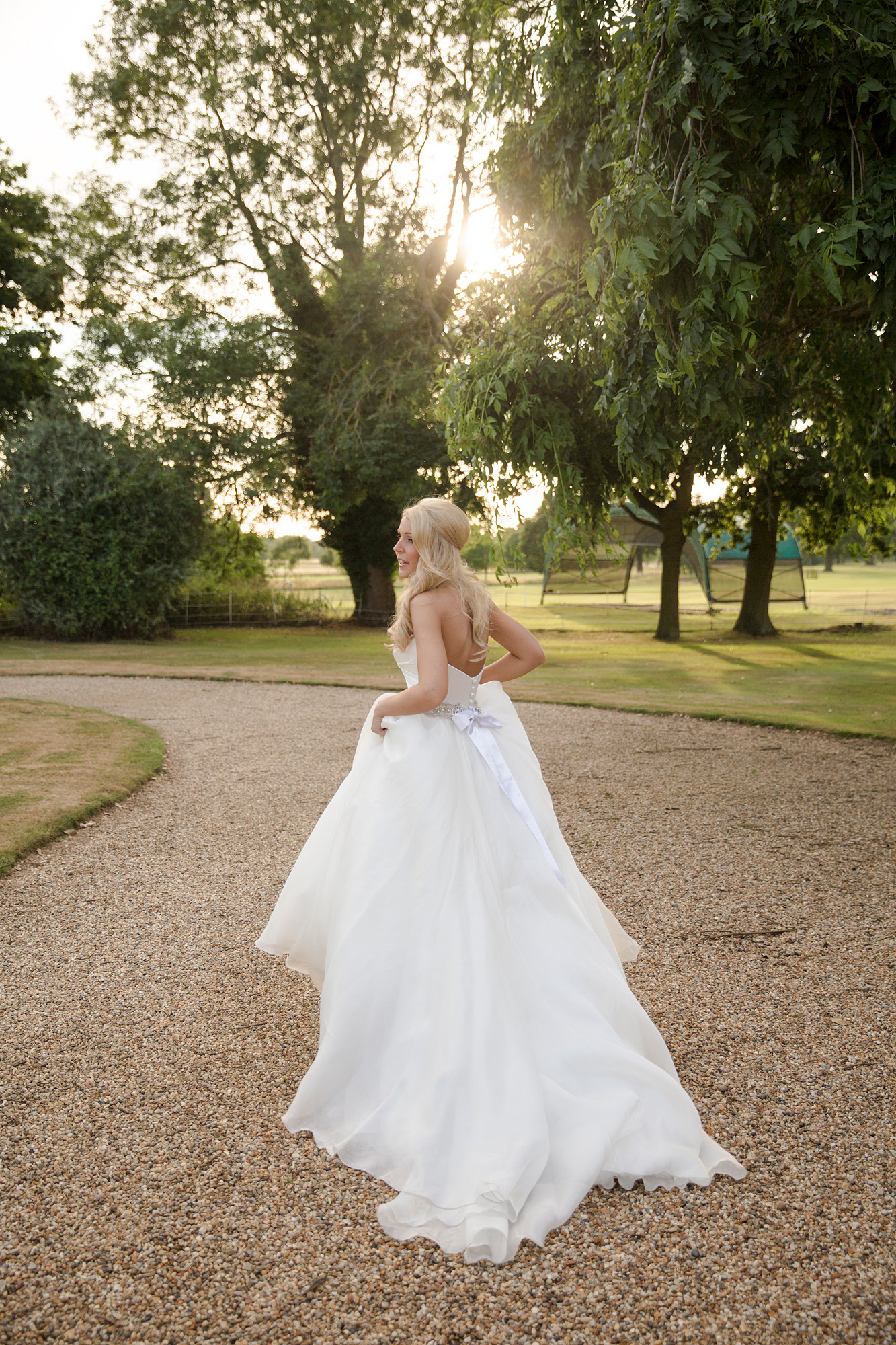 Woman in white wedding dress on gravel path in garden, sun shining through trees.