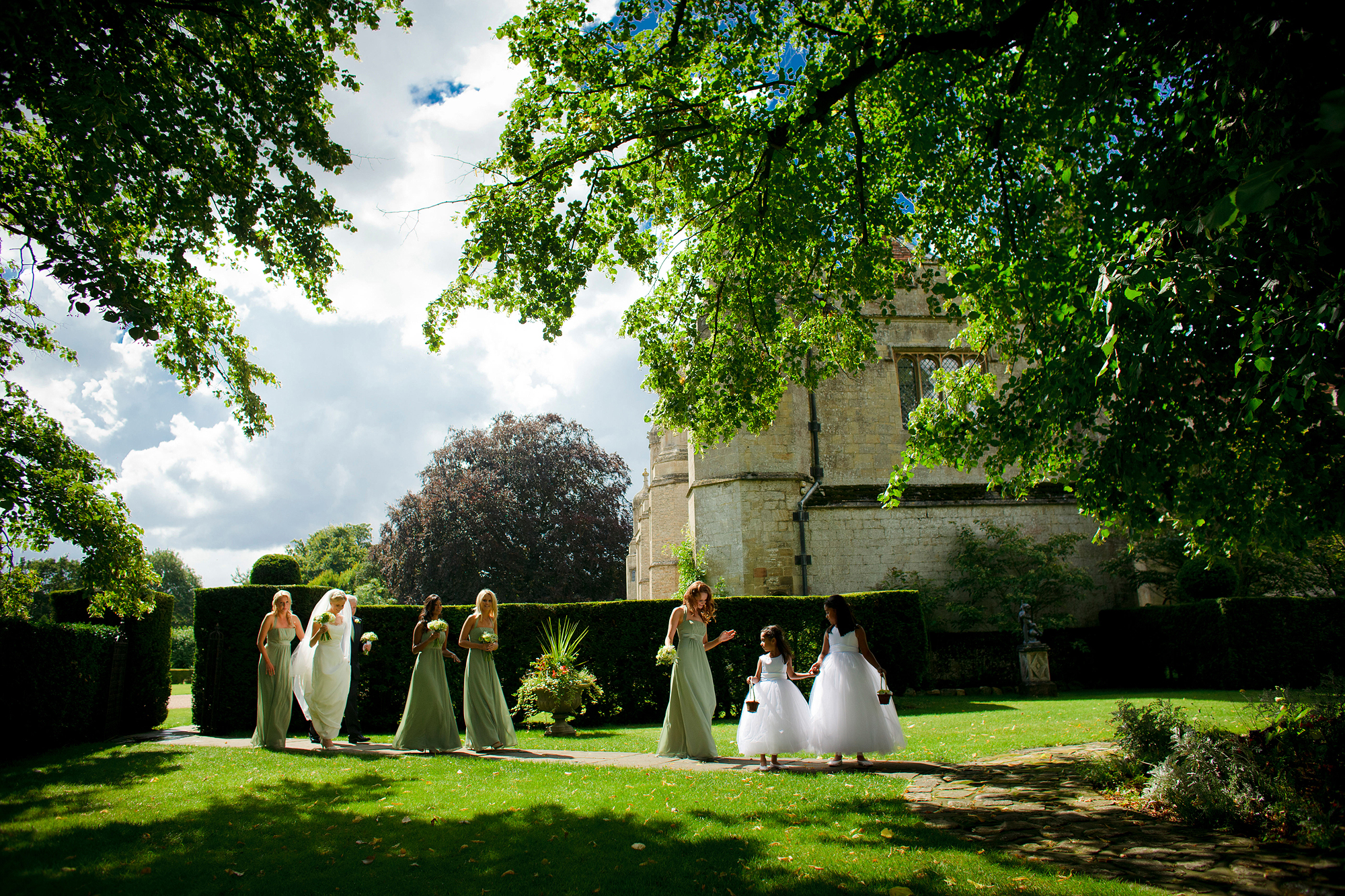 Women and children in formal attire walking through a garden near a historic stone building.
